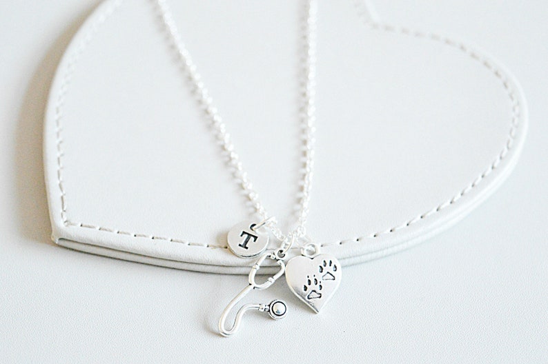 Peut inclure: Un collier argent&eacute; avec un charme de st&eacute;thoscope, un charme en forme de c&oelig;ur avec des empreintes de pattes et un charme rond avec la lettre "T". Le collier est pr&eacute;sent&eacute; sur une surface blanche en forme de c&oelig;ur.