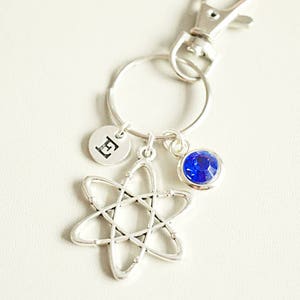 Puede incluir: Llavero de plata con un dije de cristal azul, un dije de átomo y un dije con la letra "E" en él.