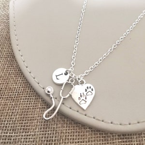 Peut inclure: Collier argent&eacute; avec un charme en forme de st&eacute;thoscope, un charme en forme de c&oelig;ur avec des empreintes de pattes et un charme rond avec la lettre "L". Le collier est pr&eacute;sent&eacute; sur une surface de couleur neutre.