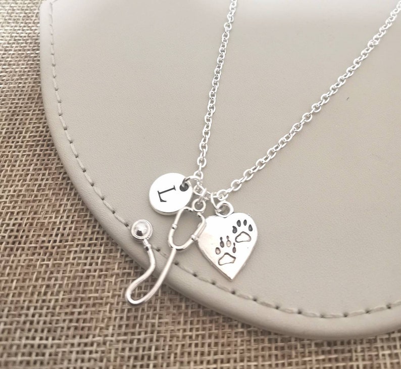 Peut inclure: Collier argent&eacute; avec un charme en forme de st&eacute;thoscope, un charme en forme de c&oelig;ur avec des empreintes de pattes et un charme rond avec la lettre "L". Le collier est pr&eacute;sent&eacute; sur une surface de couleur neutre.