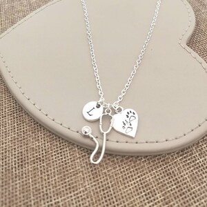Peut inclure: Un collier argent&eacute; avec un charme de st&eacute;thoscope, un charme en forme de c&oelig;ur avec des empreintes de pattes et un charme rond avec la lettre "L". Le collier est pr&eacute;sent&eacute; sur une surface en forme de c&oelig;ur gris clair.
