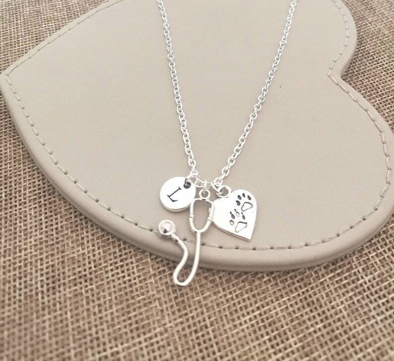 Peut inclure: Un collier argent&eacute; avec un charme de st&eacute;thoscope, un charme en forme de c&oelig;ur avec des empreintes de pattes et un charme rond avec la lettre "L". Le collier est pr&eacute;sent&eacute; sur une surface en forme de c&oelig;ur gris clair.
