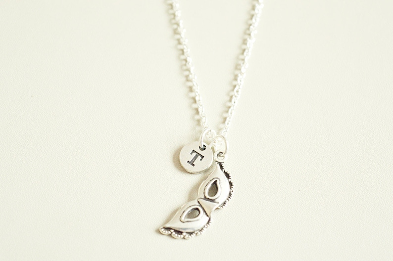 May include: A silver-toned necklace featuring a Venetian mask pendant and a small circular charm with the letter "T". The mask pendant has intricate details and a textured finish. The necklace hangs on a delicate chain.