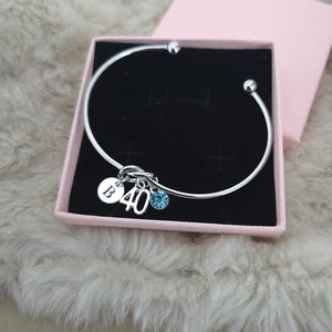 May include: A silver bangle bracelet with charms, presented in a pink gift box. The charms include a disc with the letter "B", the number "40", and a blue crystal. The bracelet is displayed on a fluffy, white surface.