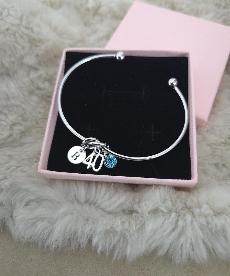 May include: A silver bangle bracelet with charms, presented in a pink gift box. The charms include a disc with the letter "B", the number "40", and a blue crystal. The bracelet is displayed on a fluffy, white surface.
