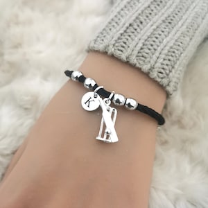 Puede incluir: Pulsera de cord&oacute;n negro con cuentas y dijes plateados. Los dijes incluyen un wicket de cricket y un disco plateado con la letra "K" en &eacute;l.