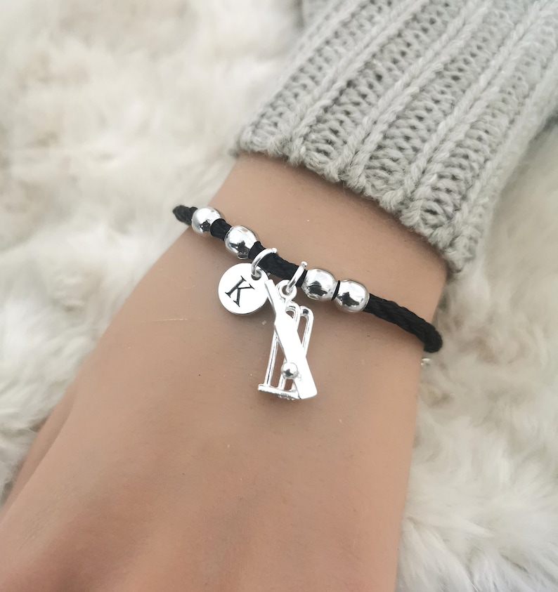 Puede incluir: Pulsera de cord&oacute;n negro con cuentas y dijes plateados. Los dijes incluyen un wicket de cricket y un disco plateado con la letra "K" en &eacute;l.