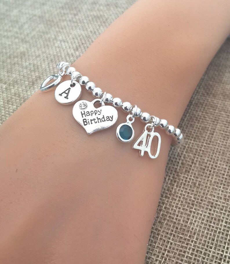May include: Silver charm bracelet with a heart charm that says "Happy Birthday", a letter "A" charm, a blue gemstone charm, and a number "40" charm.