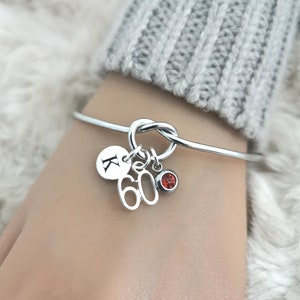 K&ouml;nnte beinhalten: Silbernes Knoten-Armband mit Charms. Die Charms umfassen einen runden Charm mit dem Buchstaben K, einen Charm mit der Zahl 60 und einen kleinen roten Edelstein-Charm.