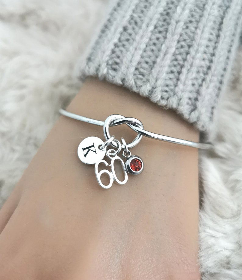 K&ouml;nnte beinhalten: Silbernes Knoten-Armband mit Charms. Die Charms umfassen einen runden Charm mit dem Buchstaben K, einen Charm mit der Zahl 60 und einen kleinen roten Edelstein-Charm.
