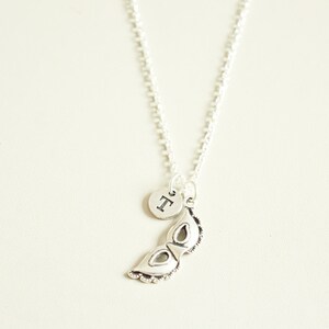 May include: Silver-toned necklace featuring a small, round pendant with a letter and a decorative mask charm. The mask charm has intricate detailing. The necklace hangs against a neutral background.
