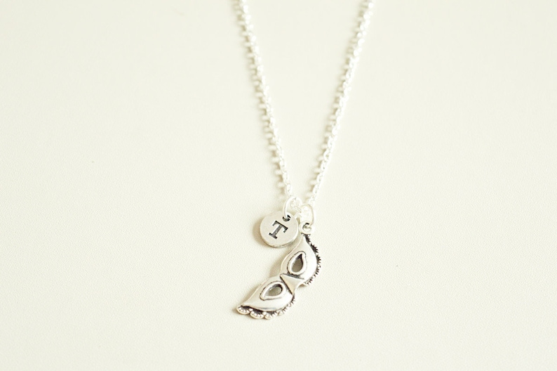 May include: Silver-toned necklace featuring a small, round pendant with a letter and a decorative mask charm. The mask charm has intricate detailing. The necklace hangs against a neutral background.