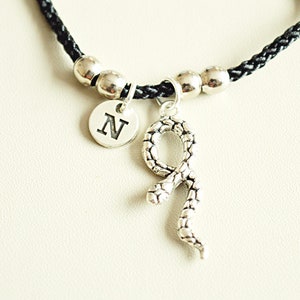 May include: A black braided cord bracelet with silver beads and a charm featuring a snake and a silver disc with the letter "N" on it.