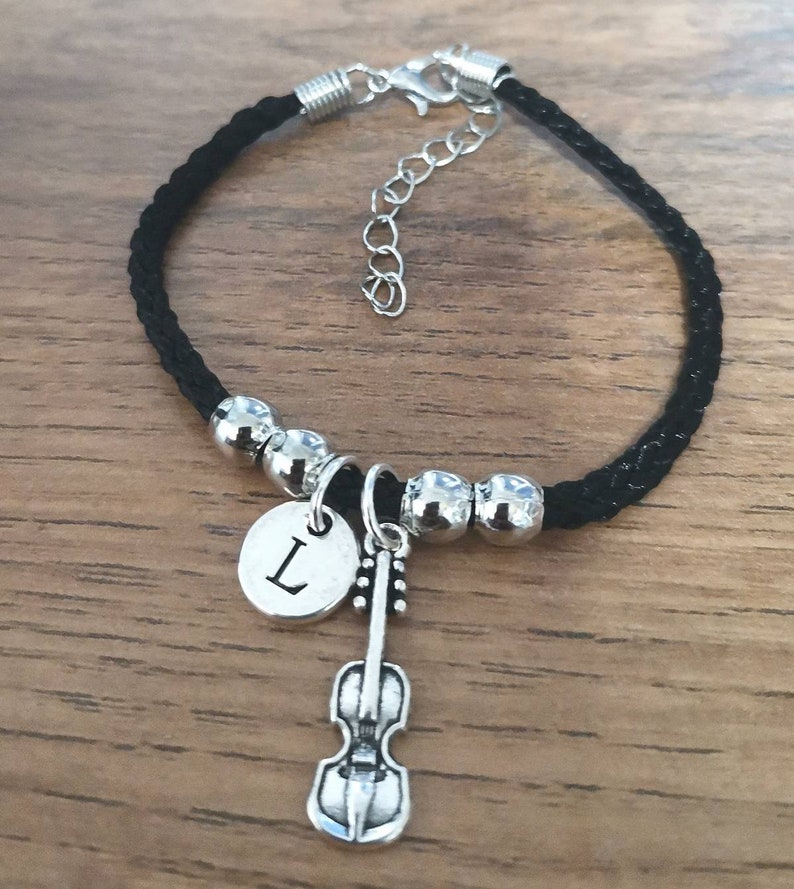 May include: Black braided cord bracelet with silver beads and a charm featuring a violin and a silver disc with the letter "L" engraved on it.