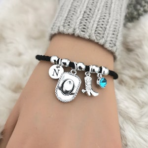 May include: A black cord bracelet with silver charms, including a cowboy hat, a cowboy boot, a turquoise blue gemstone, and a charm with the letter N.
