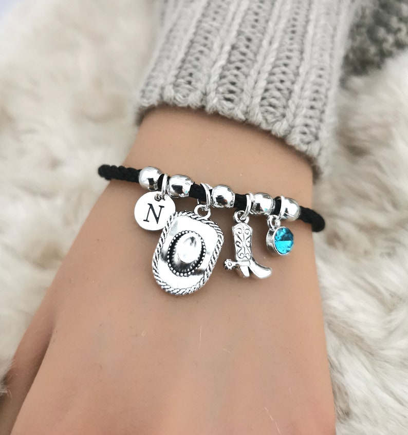 May include: A black cord bracelet with silver charms, including a cowboy hat, a cowboy boot, a turquoise blue gemstone, and a charm with the letter N.