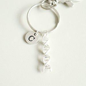 Puede incluir: Llavero de plata con un dije de doble hélice de ADN y un dije redondo con la letra "C" en él.