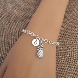 Puede incluir: Un brazalete de cadena de plata con un dije de piña y un dije de plata con la letra G.