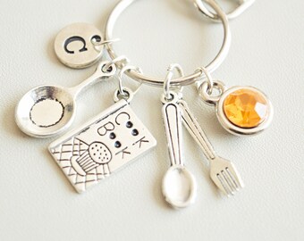 May include: Silver keychain with a cookbook charm, a spoon, a fork, a small pan, a letter 'C' charm, and an orange rhinestone.