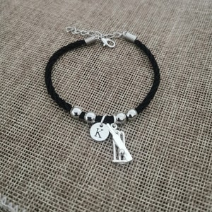 Puede incluir: Pulsera de cord&oacute;n trenzado negro con cuentas plateadas y un dije con un bate y una pelota de cricket, y un disco plateado con la letra 'K' grabada.
