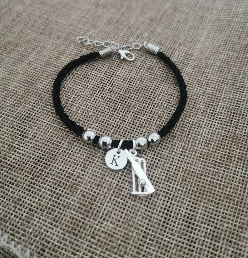 Puede incluir: Pulsera de cord&oacute;n trenzado negro con cuentas plateadas y un dije con un bate y una pelota de cricket, y un disco plateado con la letra 'K' grabada.