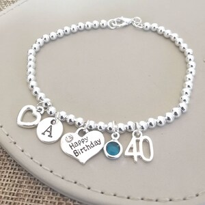 May include: Silver charm bracelet with a heart charm that says "Happy Birthday", a charm with the letter "A", a charm with the number "40", and a turquoise blue gemstone charm.