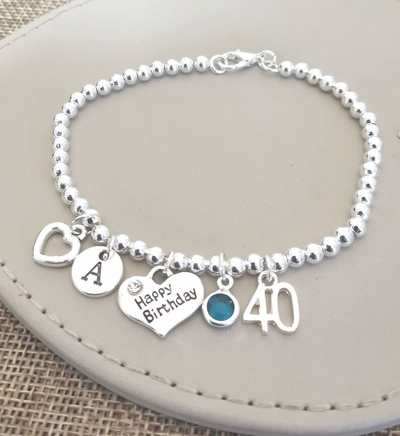 May include: Silver charm bracelet with a heart charm that says "Happy Birthday", a charm with the letter "A", a charm with the number "40", and a turquoise blue gemstone charm.