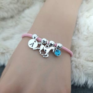 K&ouml;nnte beinhalten: Ein pinkfarbenes Armband aus Schnur mit silbernen Perlen, einem silbernen Pferdeanh&auml;nger, einem silbernen Anh&auml;nger mit dem Buchstaben A und einem hellblauen Kristallanh&auml;nger.