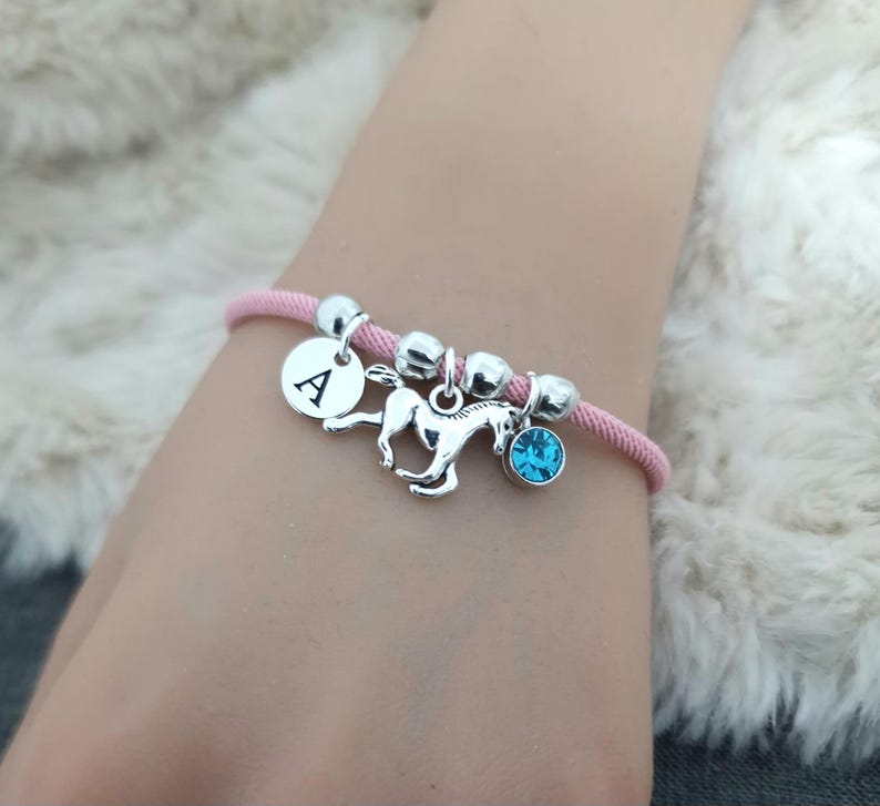 K&ouml;nnte beinhalten: Ein pinkfarbenes Armband aus Schnur mit silbernen Perlen, einem silbernen Pferdeanh&auml;nger, einem silbernen Anh&auml;nger mit dem Buchstaben A und einem hellblauen Kristallanh&auml;nger.
