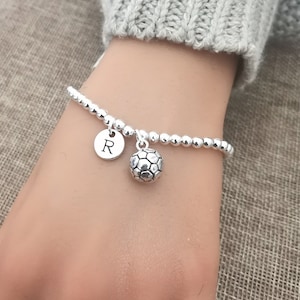 May include: Silver beaded bracelet with a soccer ball charm and a round charm with the letter "R". The bracelet is worn on a wrist, with a gray sweater in the background.