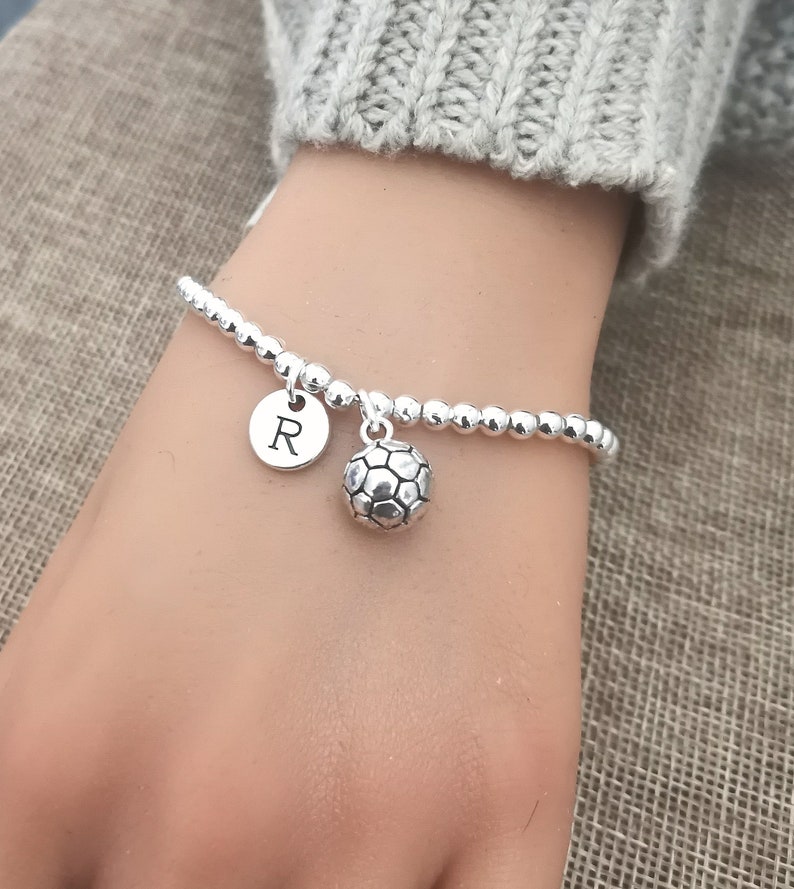 May include: Silver beaded bracelet with a soccer ball charm and a round charm with the letter "R". The bracelet is worn on a wrist, with a gray sweater in the background.
