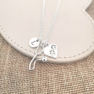 Peut inclure: Collier argent&eacute; avec un charme en forme de st&eacute;thoscope, un c&oelig;ur avec des empreintes de pattes et un charme rond avec la lettre "L". Le collier est pr&eacute;sent&eacute; sur une surface de couleur neutre.