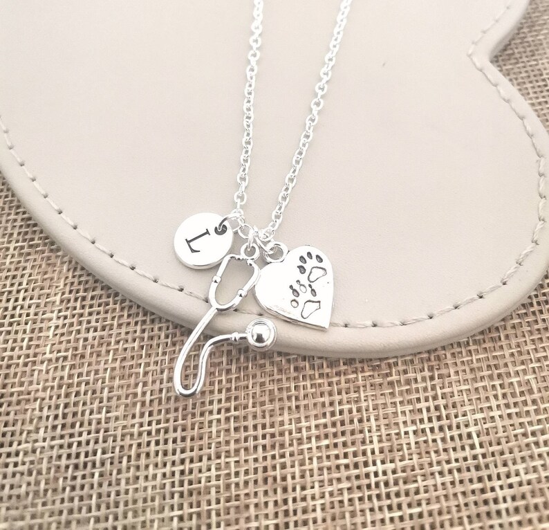 Peut inclure: Collier argent&eacute; avec un charme en forme de st&eacute;thoscope, un c&oelig;ur avec des empreintes de pattes et un charme rond avec la lettre "L". Le collier est pr&eacute;sent&eacute; sur une surface de couleur neutre.