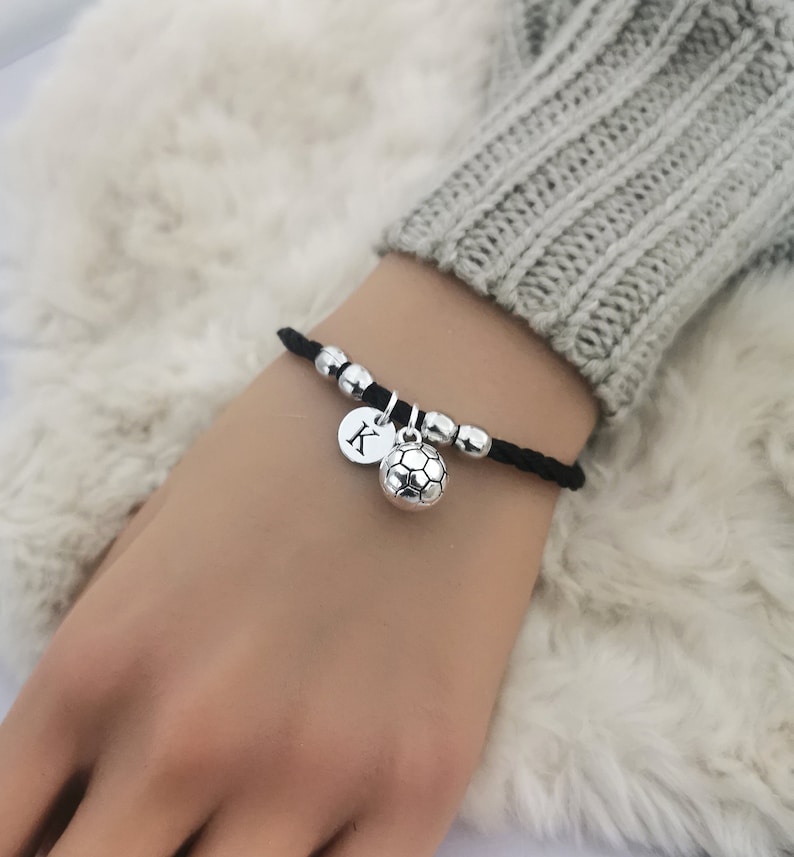 May include: Black leather bracelet with silver beads and charms. One charm is a soccer ball and the other is a silver disc with the letter K.