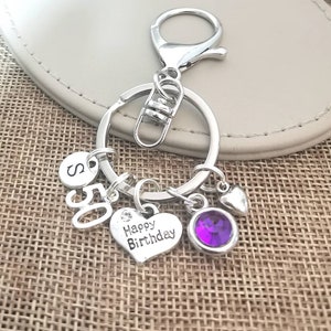 May include: Silver keychain with a purple rhinestone charm, a heart charm with the text "Happy Birthday", a charm with the number "50", and a charm with the letter "S".