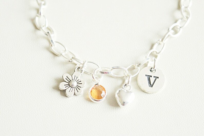 May include: A silver charm bracelet with a flower charm, a heart charm, a small amber-colored gemstone charm, and a round charm with the letter "V" engraved on it.
