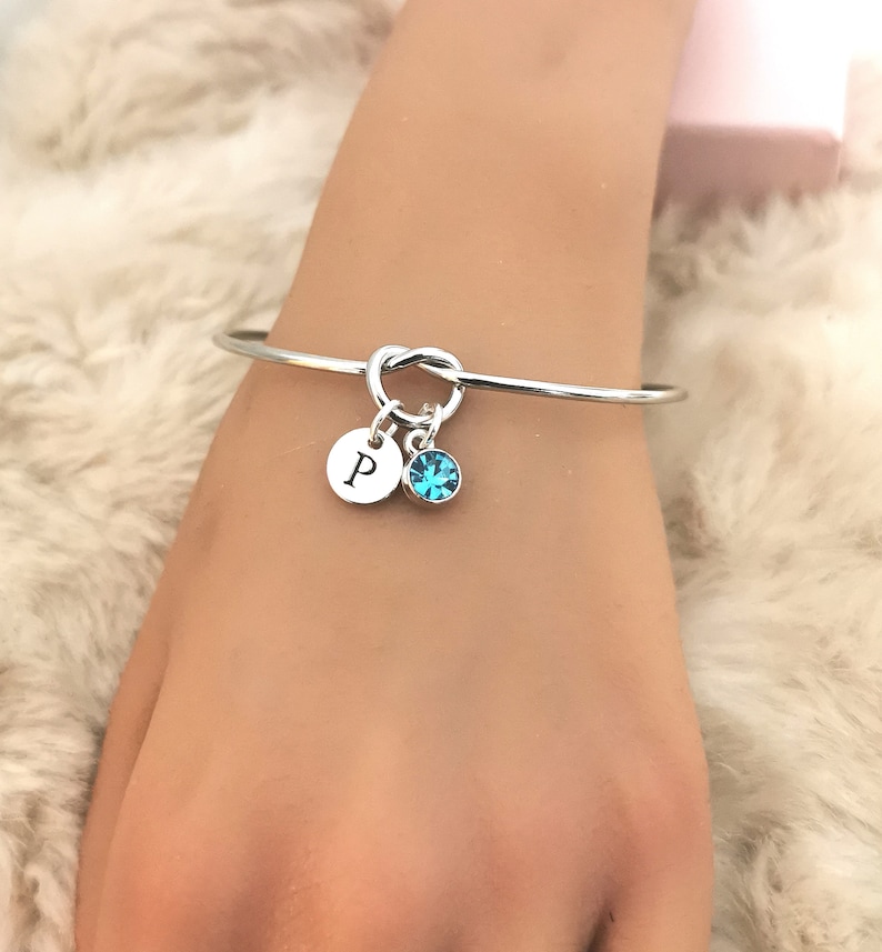 May include: Silver knot bangle bracelet with a charm featuring the letter "P" and a turquoise blue crystal.