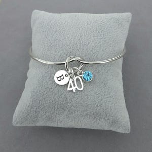 May include: A silver bangle bracelet with a knot design. It has three charms: a round charm with the letter "B", the number "40", and a blue crystal. The bracelet is displayed on a grey velvet cushion.