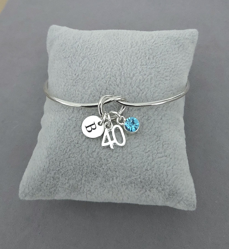 May include: A silver bangle bracelet with a knot design. It has three charms: a round charm with the letter "B", the number "40", and a blue crystal. The bracelet is displayed on a grey velvet cushion.