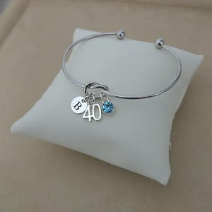 May include: Silver bangle bracelet with a knot design. It has charms with the letter "B", the number "40", and a blue gemstone. The bracelet is displayed on a white pillow.