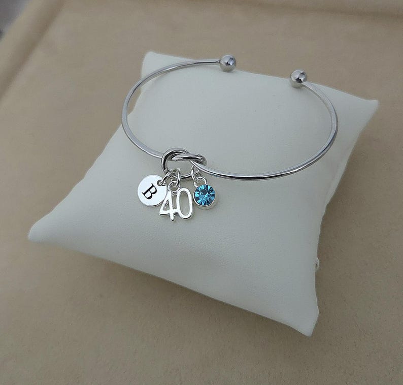 May include: Silver bangle bracelet with a knot design. It has charms with the letter "B", the number "40", and a blue gemstone. The bracelet is displayed on a white pillow.