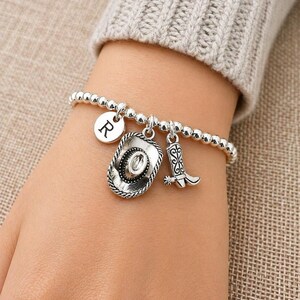 May include: Silver beaded bracelet with a cowboy hat, cowboy boot, and initial "R" charm. The bracelet is worn on a wrist, showcasing the charms' details. The bracelet is a western-themed accessory.