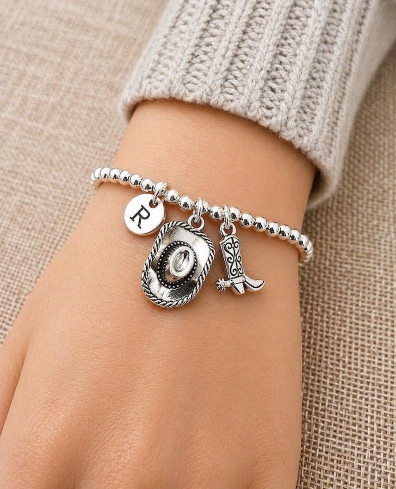 May include: Silver beaded bracelet with a cowboy hat, cowboy boot, and initial "R" charm. The bracelet is worn on a wrist, showcasing the charms' details. The bracelet is a western-themed accessory.
