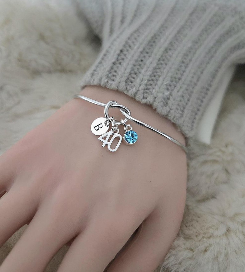 May include: A silver bangle bracelet with three charms. One charm is a round disc with the letter "B", another is the number "40", and the third is a blue crystal. The bracelet is worn on a wrist.