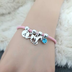 K&ouml;nnte beinhalten: Ein pinkfarbenes Armband aus Kordel mit silbernen Perlen, einem silbernen Pferdeanh&auml;nger, einem silbernen Anh&auml;nger mit dem Buchstaben A und einem blauen Kristallanh&auml;nger.