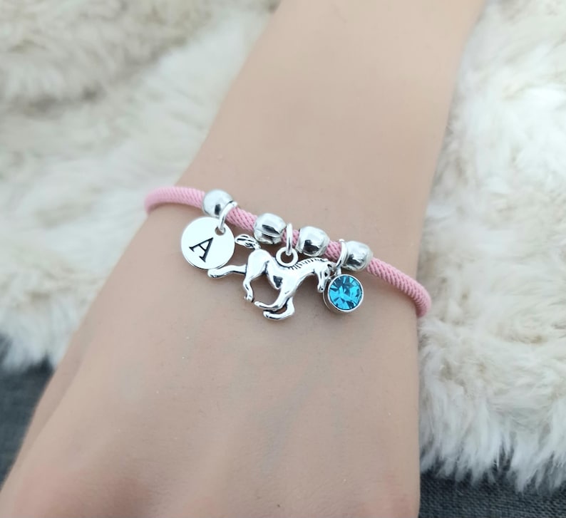 K&ouml;nnte beinhalten: Ein pinkfarbenes Armband aus Kordel mit silbernen Perlen, einem silbernen Pferdeanh&auml;nger, einem silbernen Anh&auml;nger mit dem Buchstaben A und einem blauen Kristallanh&auml;nger.