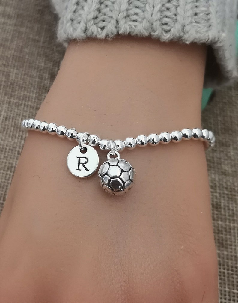 May include: Silver beaded bracelet with a soccer ball charm and a round charm with the letter "R". The bracelet is on a wrist, with a gray sweater sleeve in the background.