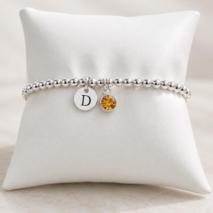 Könnte beinhalten: Ein elastisches Armband aus glänzenden silberfarbenen Perlen, verziert mit einem kleinen runden Anhänger, der mit dem Buchstaben D graviert ist, sowie einem weiteren Anhänger mit einem runden, bernsteinfarbenen Schmuckstein.
