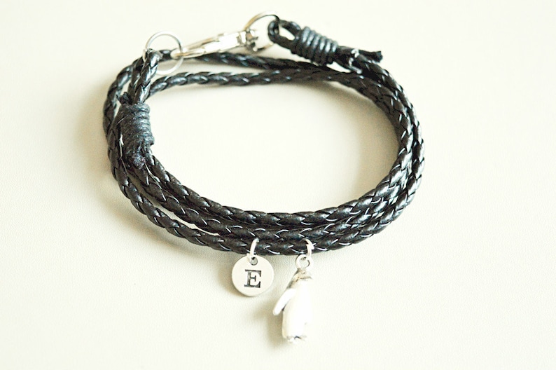 Op de afbeelding: Een zwarte leren gevlochten armband met een zilveren bedel met de letter 'E' erop en een zilveren pingu&iuml;nbedel.