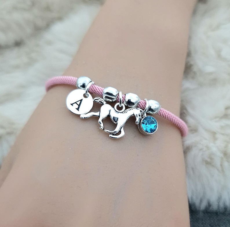 K&ouml;nnte beinhalten: Rosafarbenes Kordelarmband mit silbernen Perlen, einem silbernen Pferdeanh&auml;nger, einem silbernen Anh&auml;nger mit dem Buchstaben A und einem hellblauen Kristallanh&auml;nger.